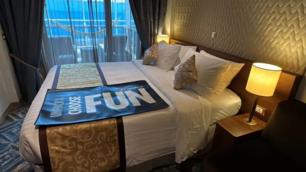 The hotel room, reminiscent of Carnival Venezia cabins, boasts a large bed adorned with white and gold linens. A "Choose Fun" banner graces the foot of the bed. Two lamps softly illuminate the space, while glass doors reveal a balcony offering a picturesque view in the background.