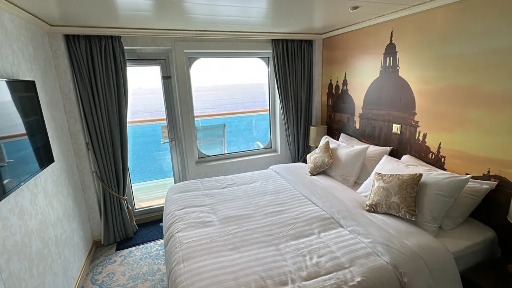 A cozy Carnival Venezia cabin features a large bed with decorative pillows and a mural of a domed building. The space includes a window and door leading to a balcony with an ocean view, while a flat-screen TV is mounted on the wall.