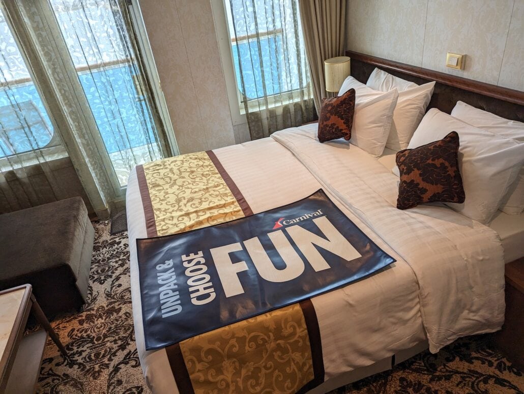 A neatly made bed in a Carnival Venezia cabin features white linens and decorative pillows. The large window offers a stunning sea view, framed by ivory curtains. A banner with "Unpack & Choose Fun" and the Carnival logo completes this inviting cruise ship retreat for ultimate relaxation.