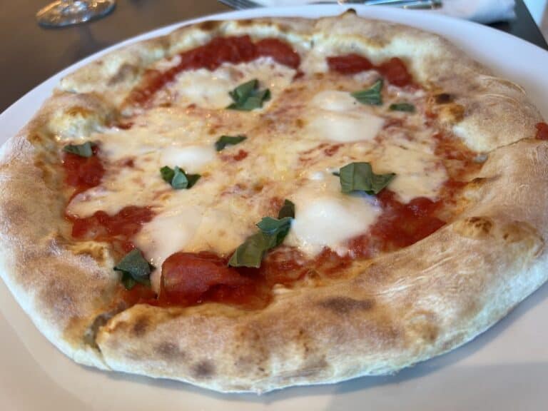 A close-up image of a Margherita pizza on a white plate, reminiscent of the delicious cuisine offered by Princess Cruises. The pizza boasts a golden-brown crust, rich tomato sauce, melted mozzarella cheese, and fresh basil leaves. A fork and napkin are visible in the background.