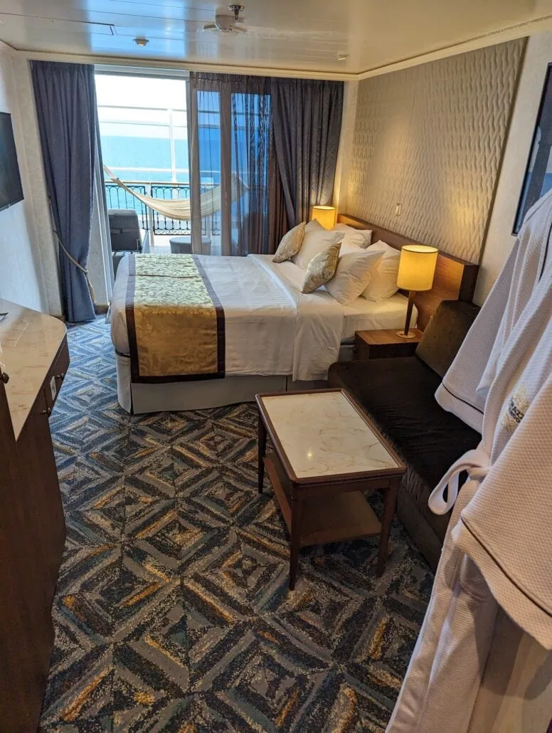 A hotel room with a double bed featuring gold and white bedding, two bedside lamps, a sofa, and a small table reminiscent of the elegance seen in Carnival Venezia cabins. A wall-mounted TV is on the left, while sliding glass doors open to a balcony with an ocean view. Bathrobes hang on the right.