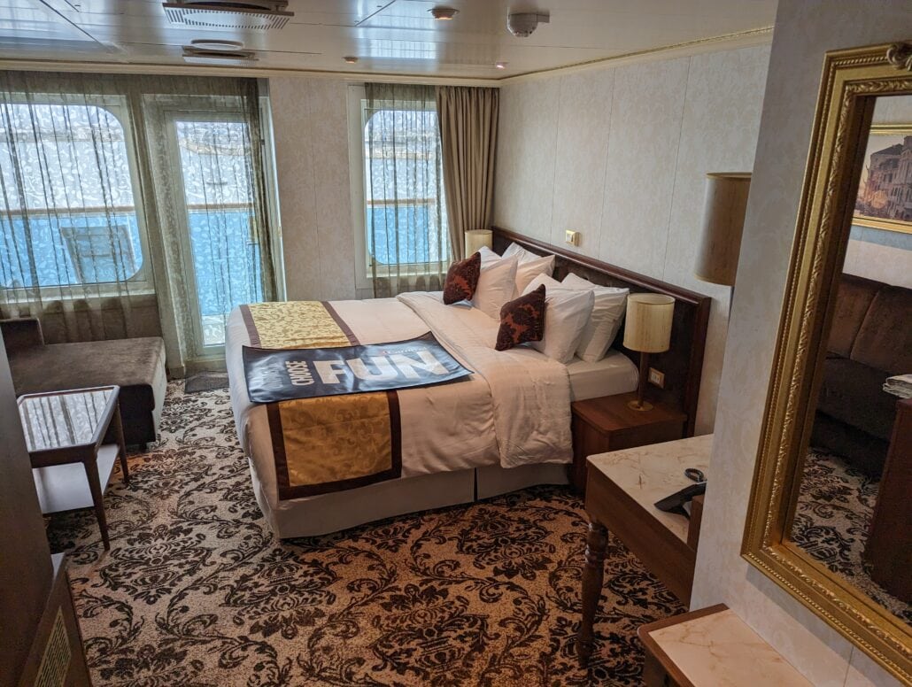The hotel room, reminiscent of Carnival Venezia cabins, boasts a large bed adorned with white linens and brown pillows. Ornate carpet and patterned curtains complement the ocean-view window, while a small couch, mirror, and nightstand with a lamp complete this inviting sanctuary.