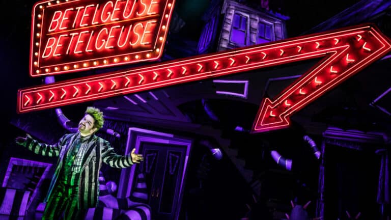 A performer dressed in black and white striped clothing stands on a stage under a large neon sign displaying "BETELGEUSE" three times. The stage, part of Norwegian Viva Entertainment, is decorated with vibrant, surreal set pieces, including an arrow pointing towards the performer.