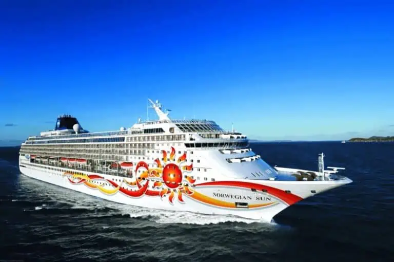 The image shows a large cruise ship named Norwegian Sun sailing on the ocean with colorful sun-themed artwork on its white hull. With a clear, blue sky suggesting a sunny day, this picturesque scene marks NCL's return to Baltimore. Small waves ripple in the water, adding to the serene setting.