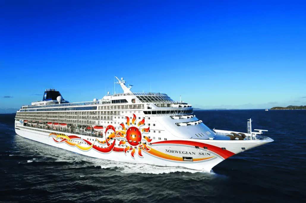 The image shows a large cruise ship named Norwegian Sun sailing on the ocean with colorful sun-themed artwork on its white hull. With a clear, blue sky suggesting a sunny day, this picturesque scene marks NCL's return to Baltimore. Small waves ripple in the water, adding to the serene setting.