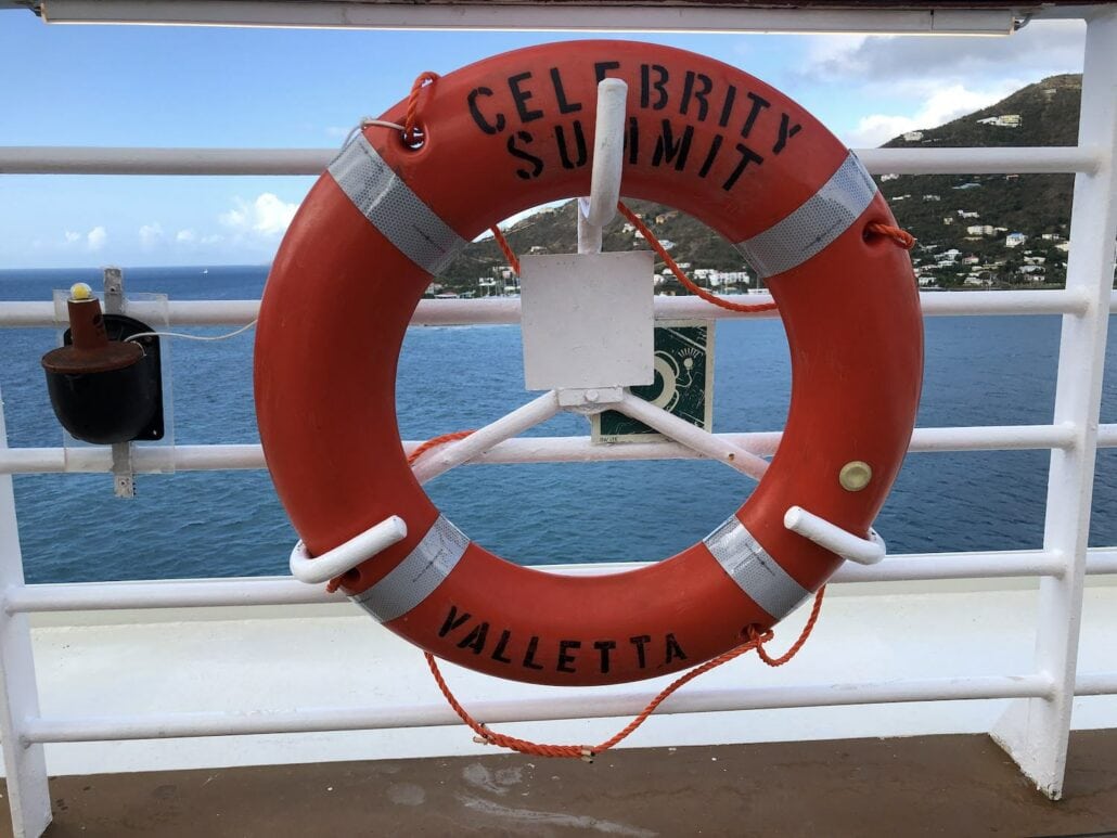 A red lifebuoy emblazoned with "Celebrity Summit" and "Valletta" is secured to a white railing on the ship, overlooking the vast ocean. Nearby, a small reflective sign and a distinctive black and red box are mounted. In the background, rolling hills gracefully enhance the vista.