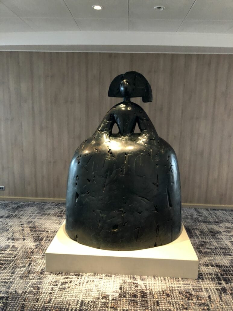 A large, abstract bronze sculpture with smooth, rounded forms is displayed indoors on a white pedestal, reminiscent of the elegance found aboard the Celebrity Summit. The background features a wooden-panel wall and a textured carpeted floor, creating an air of sophisticated luxury.