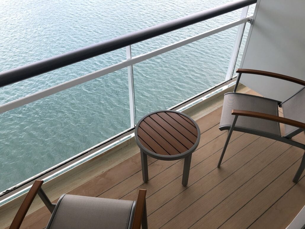 A small balcony on the Celebrity Summit overlooks a body of water, featuring two chairs and a round wooden-top table. The wood-paneled floor complements the glass railing, offering an unobstructed view of the serene waters below.