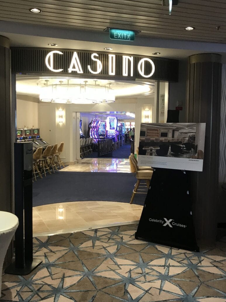 The entrance to the casino on the Celebrity Summit cruise ship features a sign above the doorway. Inside, slot machines and gaming tables beckon players. A promotional sign with "Celebrity Cruises" is placed near the entrance, while a decorative ceiling design adds an elegant touch.