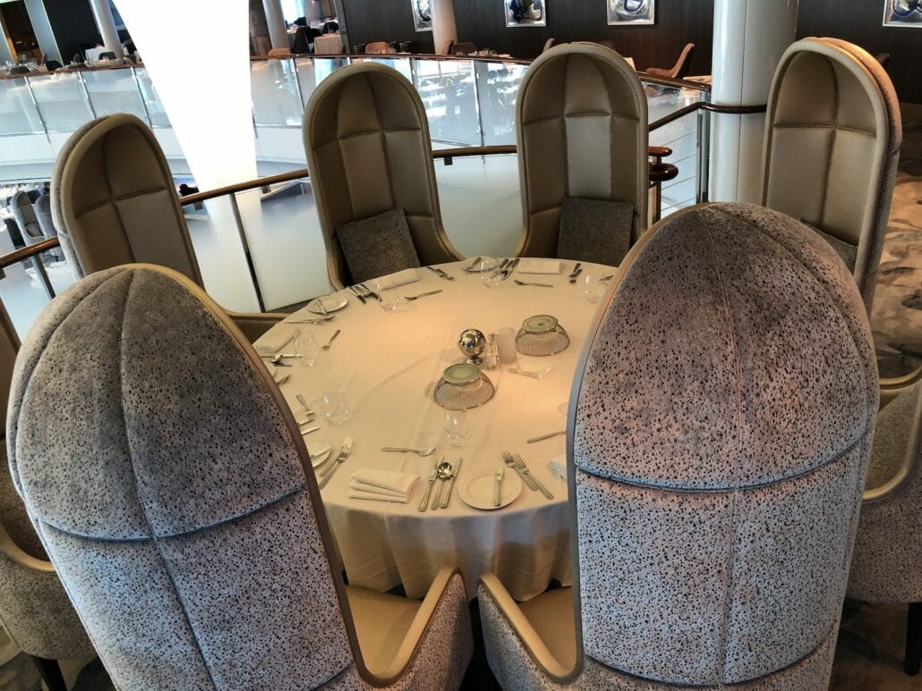 A round dining table with a white tablecloth is set for six, featuring high-backed, cushioned chairs. The arrangement includes plates, cutlery, and small domed covers, offering a luxurious dining experience akin to that on the Celebrity Summit in this modern restaurant with glass railings.