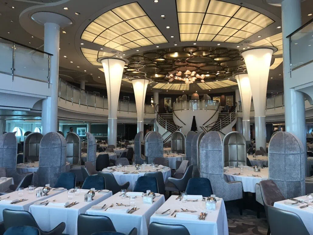 A spacious and elegant dining area on the Celebrity Summit features white tablecloth-covered tables, blue and gray chairs, and distinctive gray booths. With a high ceiling, modern lighting, and a mezzanine level, it creates a luxurious atmosphere perfect for an exceptional dining experience.