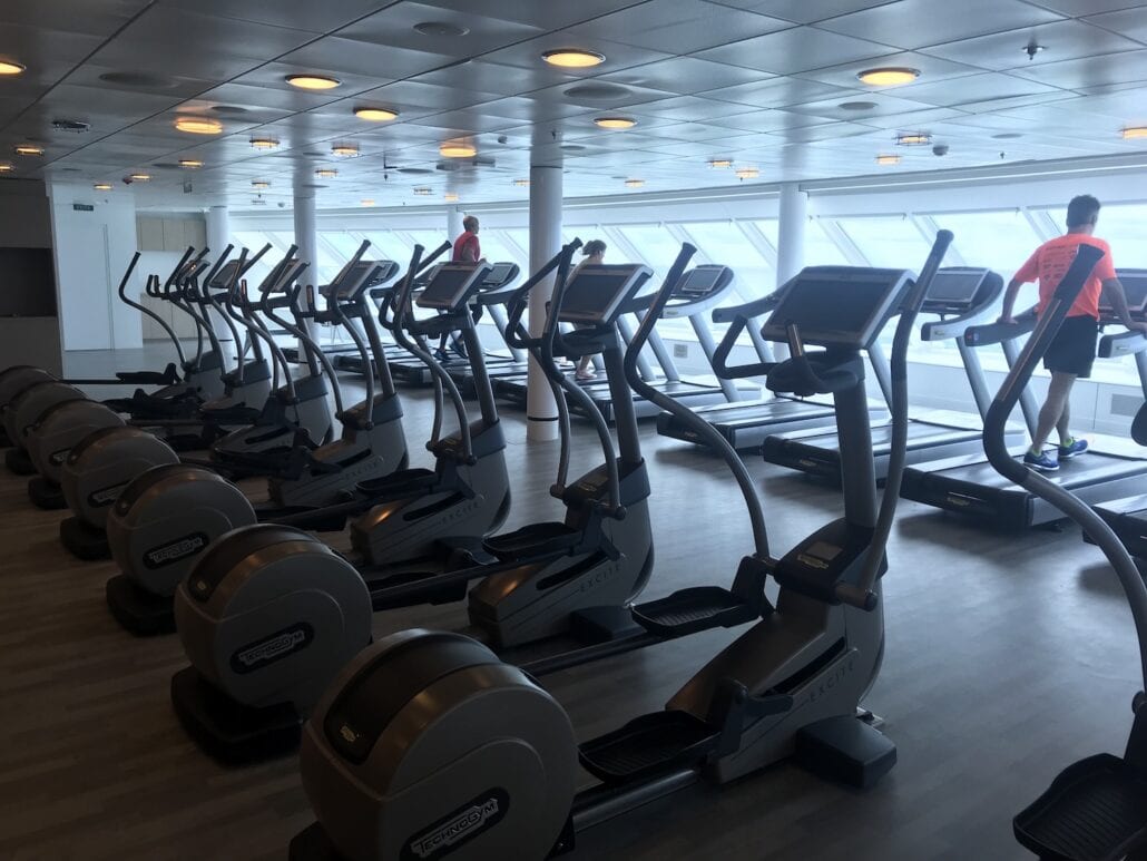 A row of treadmills and elliptical machines shines under the recessed lighting in a well-lit gym reminiscent of a fitness deck on the Celebrity Summit. Two people are exercising with determination, framed by wide windows that reveal a beautiful ocean view. The wood-like floor adds to this luxurious atmosphere.