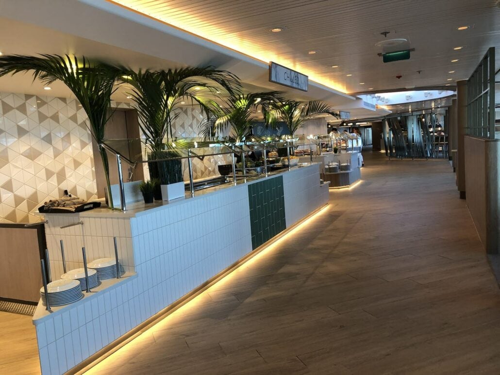 A brightly lit buffet area with a modern design, reminiscent of the Celebrity Summit, features a long counter with plates and food trays, flanked by decorative palm plants. The space boasts a tiled wall and wooden floor, seamlessly leading to an open seating area.