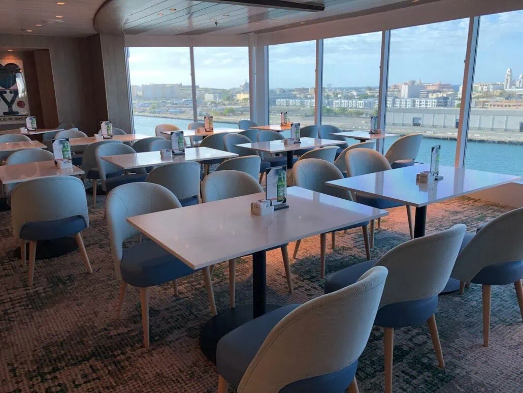 A modern dining area aboard the Celebrity Summit features empty tables and chairs in soft blue tones. Large windows offer a stunning view of the cityscape and harbor. The tables are set with menus and condiment holders, while a carpeted floor with a geometric pattern adds to the stylish ambiance.