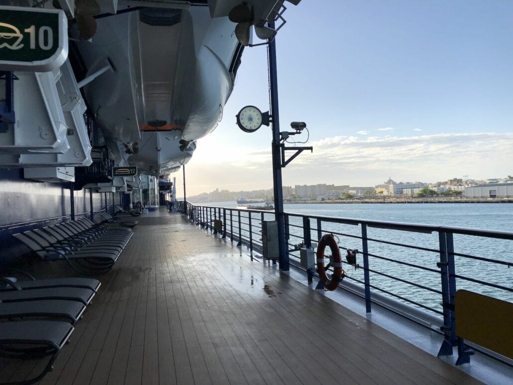 View of the Celebrity Summit cruise ship deck with lifeboats and lounge chairs. A clock and a life preserver are mounted on the railing. The background features a calm sea and a distant skyline under a clear sky.
