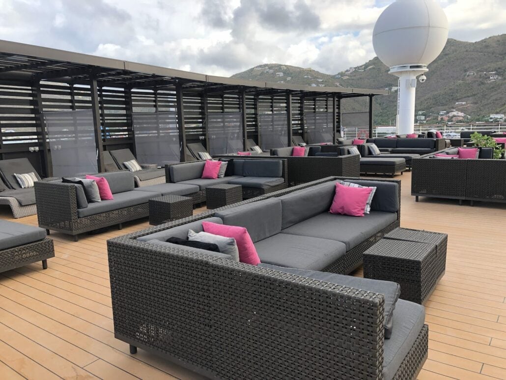 The Celebrity Summit's outdoor seating area features a wooden deck adorned with gray wicker couches and pink accent pillows. A large white sphere adds a touch of intrigue, set against a backdrop of rolling hills under a cloudy sky.