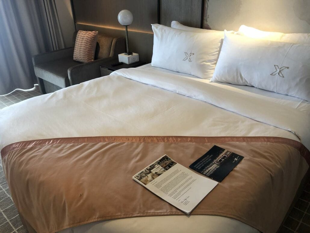 A neatly made hotel bed with white linens and two pillows marked with an "X" evokes a Celebrity Summit suite. A beige throw graces the foot, while a magazine and document rest on top. In the background, a gray sofa and lamp complete this serene setting.