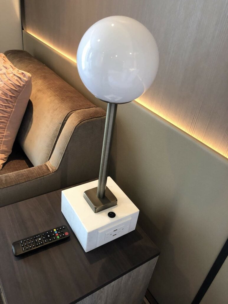 A modern lamp with a round white shade rests on a square base with power outlets, reminiscent of the sleek designs you might find aboard the Celebrity Summit. It's situated on a dark wooden side table, hosting a remote control next to a beige couch adorned with a pink cushion. Warm lighting graces the wall in the background.