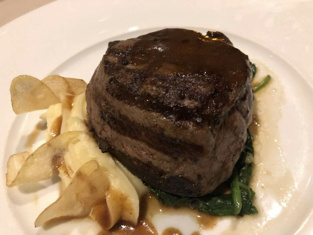 A cooked filet mignon sits on a white plate aboard the Celebrity Summit, garnished with a dark sauce. It rests on a bed of spinach and mashed potatoes, accompanied by thinly sliced potato chips.