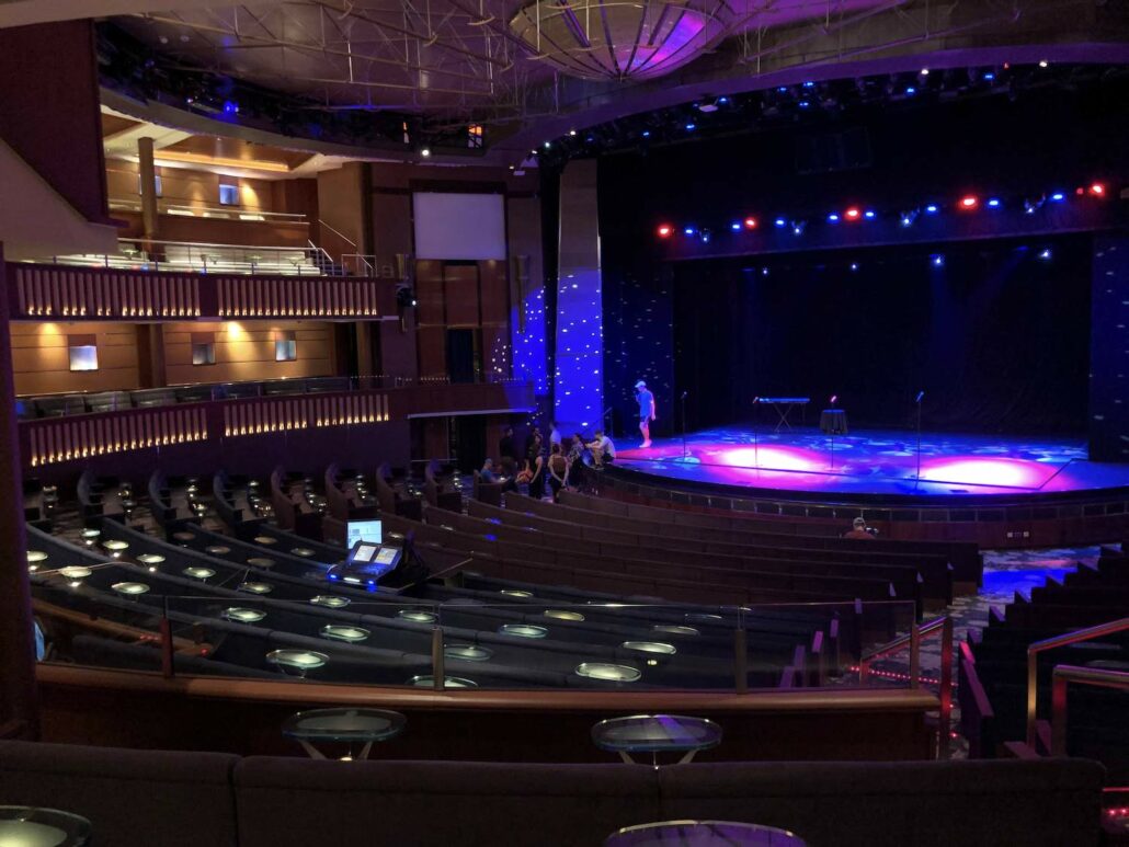 An empty auditorium with tiered seating and tables faces a stage illuminated by spotlighting, reminiscent of a Celebrity Summit. The stage, equipped with a microphone and keyboard stand, is enhanced by blue and red lights. Balcony seating runs along the sides and rear of the venue.