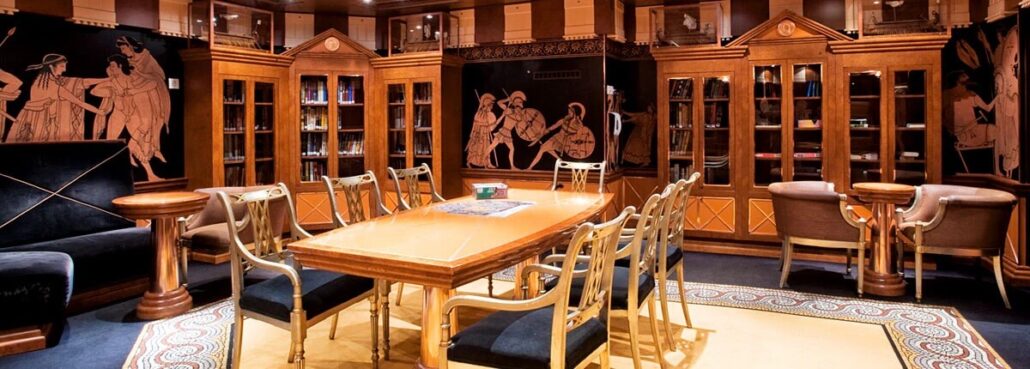 A spacious room on the cruise ship featuring a large wooden table surrounded by chairs. The walls are lined with bookshelves, adorned with classical-style murals depicting figures in historical attire. Soft lighting creates a warm atmosphere, perfect for those seeking quiet moments amidst the carnival spirit.