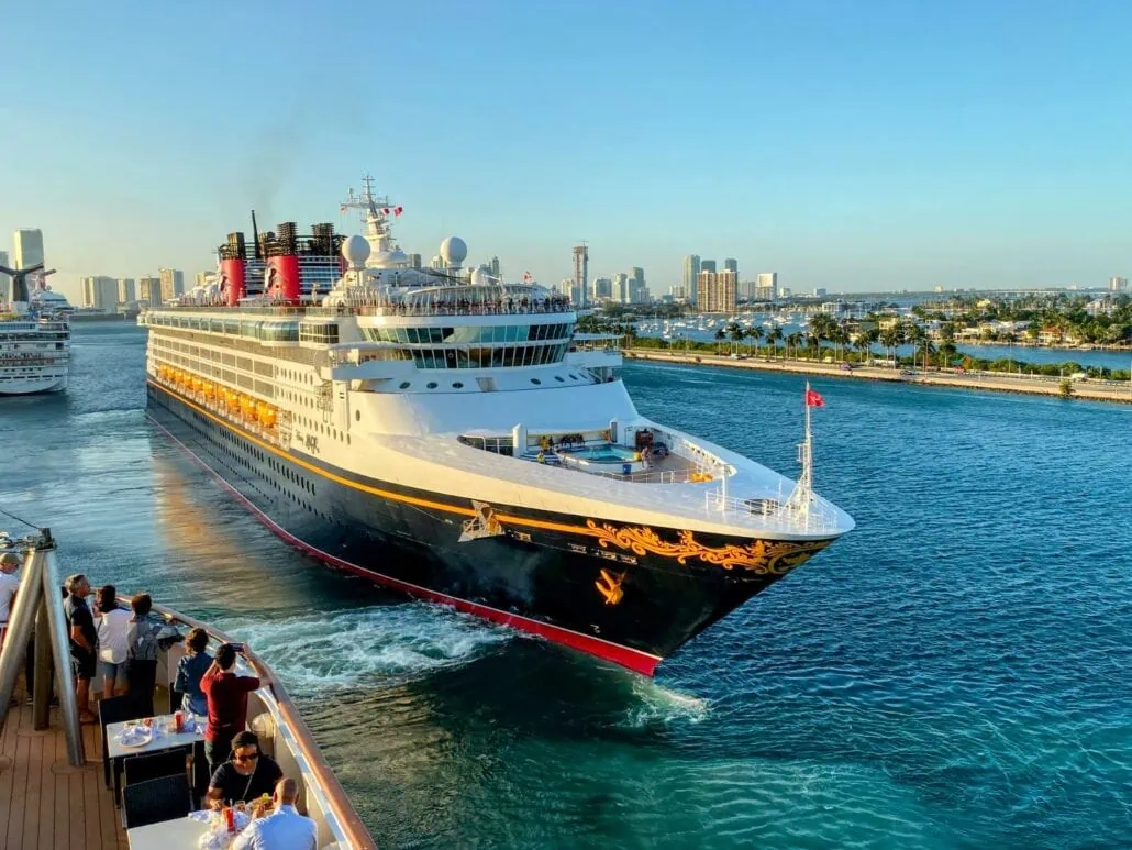 A large Disney cruise ship sails in a sunny coastal city port. Passengers on another vessel watch and take pictures. Buildings and palm trees are visible in the background.