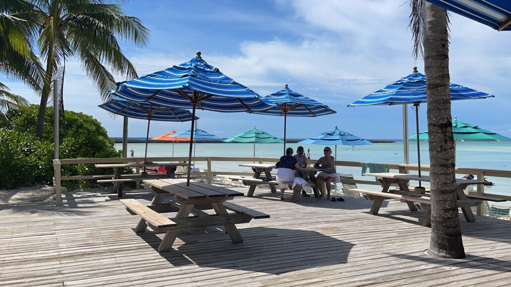 A small group of people sit at a wooden picnic table under blue-striped umbrellas on a wooden deck by the beach at Castaway Cay. There are palm trees and more umbrellas in the background, with the ocean and a clear blue sky visible.
