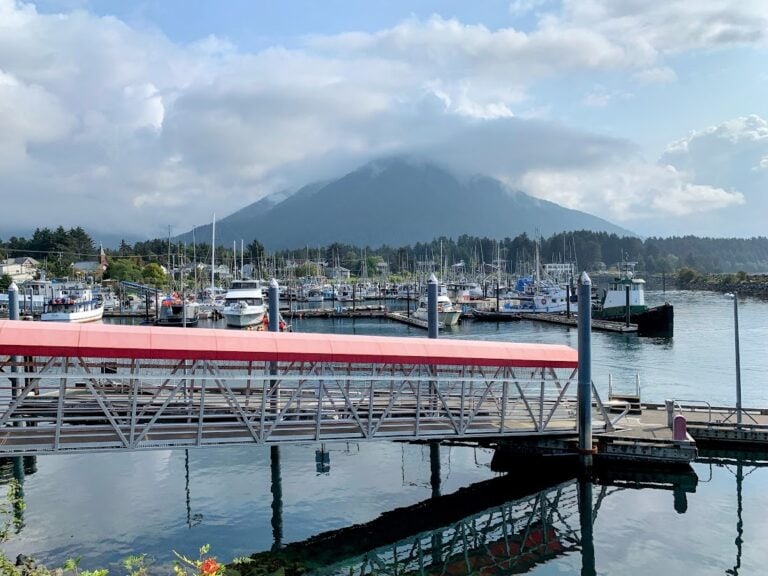 A marina with several boats docked along the piers showcases the serene charm of an Alaska port. A bridge with a red canopy crosses over calm water, while in the background, a cloud-kissed mountain rises behind the town under a partly cloudy sky.