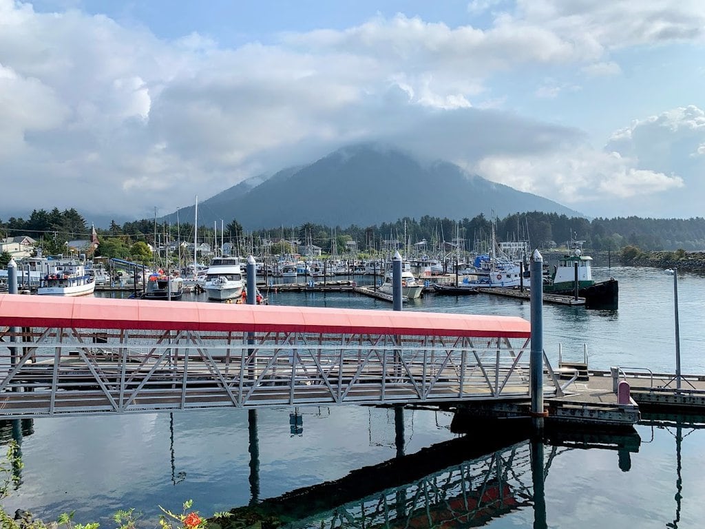 A marina with several boats docked along the piers showcases the serene charm of an Alaska port. A bridge with a red canopy crosses over calm water, while in the background, a cloud-kissed mountain rises behind the town under a partly cloudy sky.