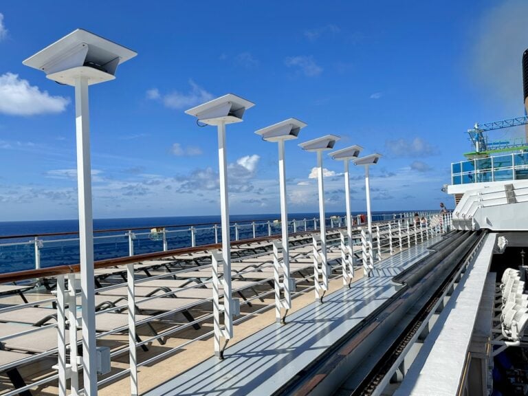 The image shows a cruise ship deck with white lounge chairs lined up under a clear blue sky. Starlink high-speed internet ensures guests stay connected, as a row of white lamp posts is positioned along the railing, with the ocean visible in the background.