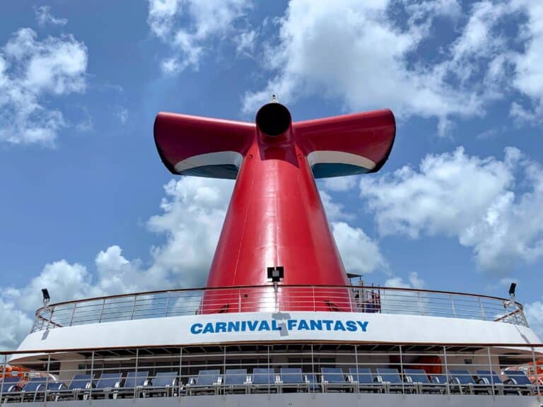 A large red funnel with a white and blue accent stands atop the Carnival Fantasy cruise ship—one of several carnival ships sold—against a partly cloudy sky. The ship’s name, “CARNIVAL FANTASY,” appears in blue letters on the deck below.