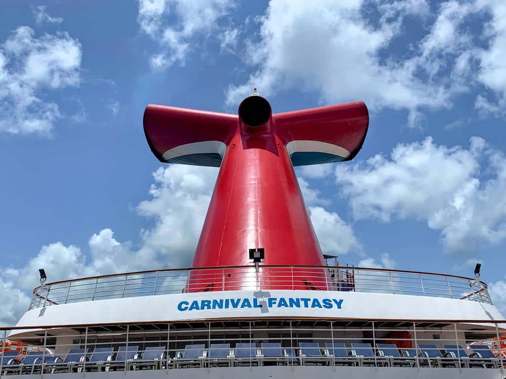 A large red funnel with a white and blue accent stands atop the Carnival Fantasy cruise ship—one of several carnival ships sold—against a partly cloudy sky. The ship’s name, “CARNIVAL FANTASY,” appears in blue letters on the deck below.