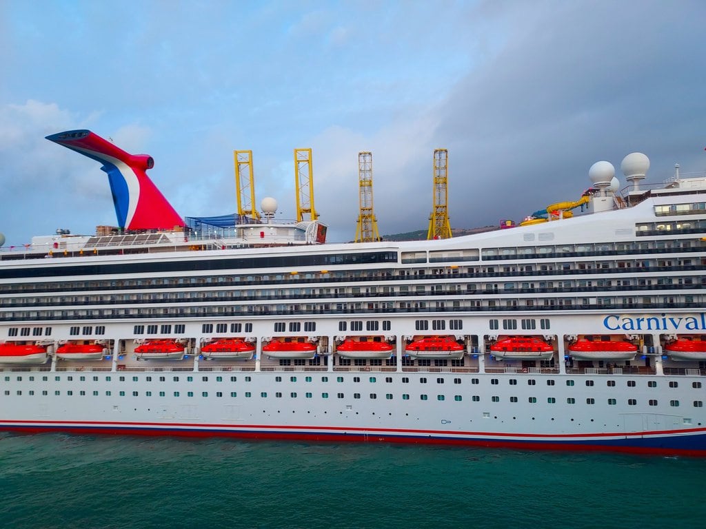 A large white cruise ship named Carnival Freedom displays the word "Carnival" on its side. The ship boasts multiple decks, with red lifeboats and yellow structures visible on the top deck. The sky appears cloudy, and the ship is situated in calm greenish water, showcasing its recent makeover.