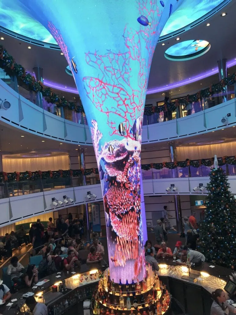 A vibrant interior scene aboard the Carnival Vista features a large, colorful digital display shaped like a column, depicting underwater imagery. Guests are seated around a circular bar beneath it. Holiday decorations, including a Christmas tree, add to the festive atmosphere of this cruise review setting.