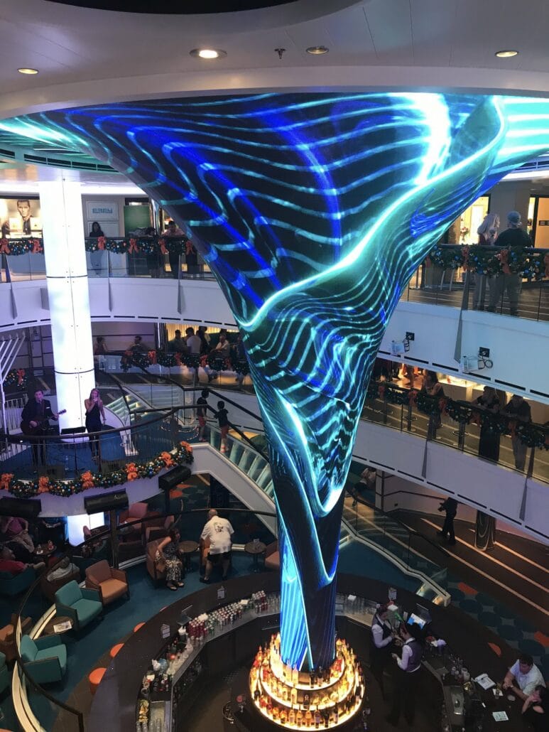 A vibrant atrium aboard the Carnival Vista features a large, swirling LED display resembling a vortex, with mesmerizing blue and green patterns. People gather on different levels with a circular bar at the base of this stunning attraction, creating an unforgettable cruise experience.