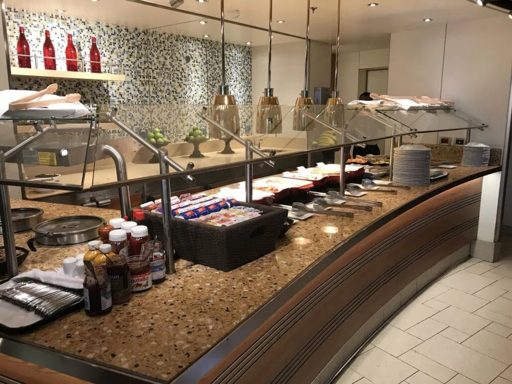 A buffet counter in the restaurant aboard the Carnival Vista cruise is laden with assorted foods, condiments, and dishes. Trays brim with delicacies, packets of zesty sauces, and neatly stacked plates. The decorative tiled wall adds charm under the warm glow of overhead lights.