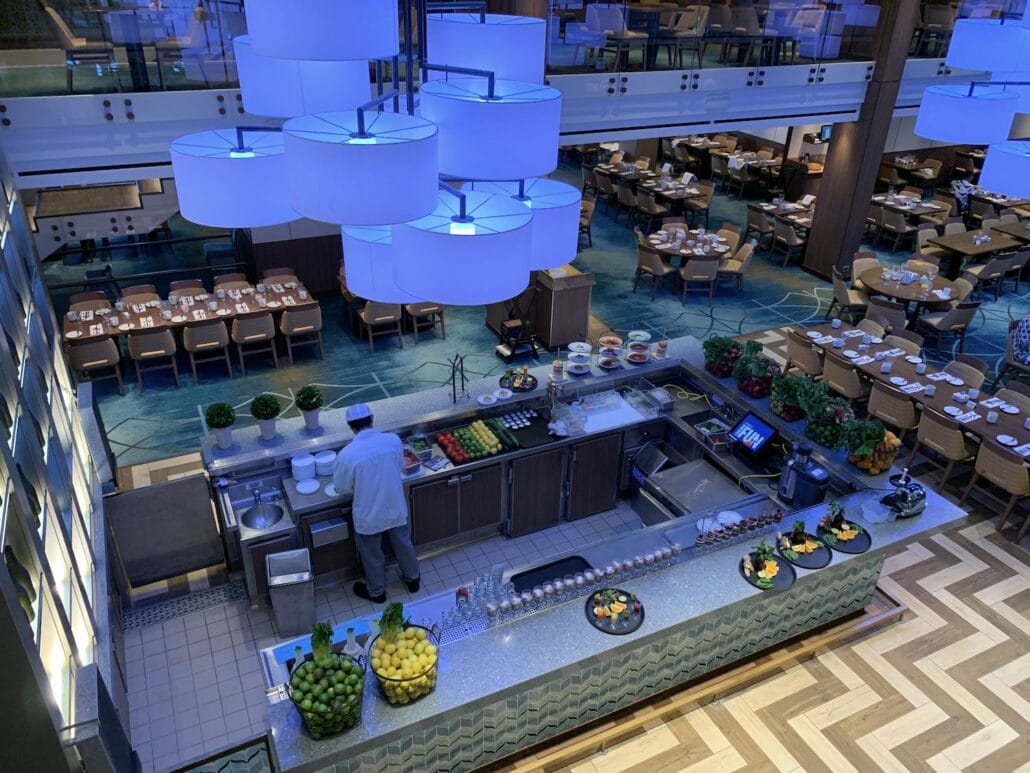 A high-angle view of a Carnival Vista cruise buffet showcases a counter brimming with fruits, ingredients, and plates. A chef in a white coat skillfully prepares dishes. In the background, the dining area is neatly arranged with tables and chairs, illuminated by large blue ceiling lights.
