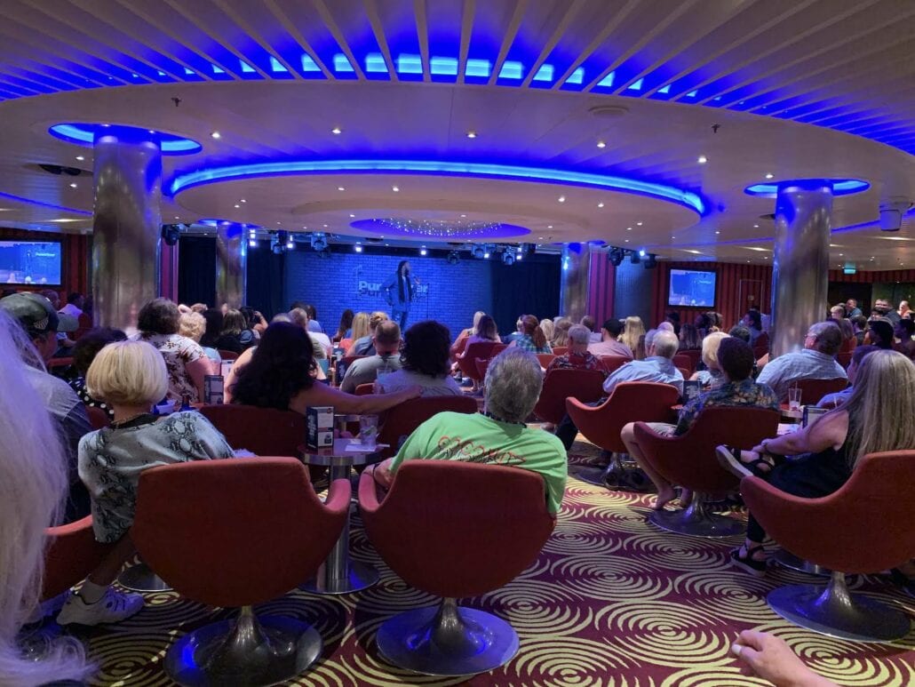 A crowd sitting in red chairs faces a stage where a person delivers stand-up comedy. With a circular ceiling illuminated in blue and TV screens showcasing the performance, it's like an entertaining night aboard a Carnival Vista cruise.