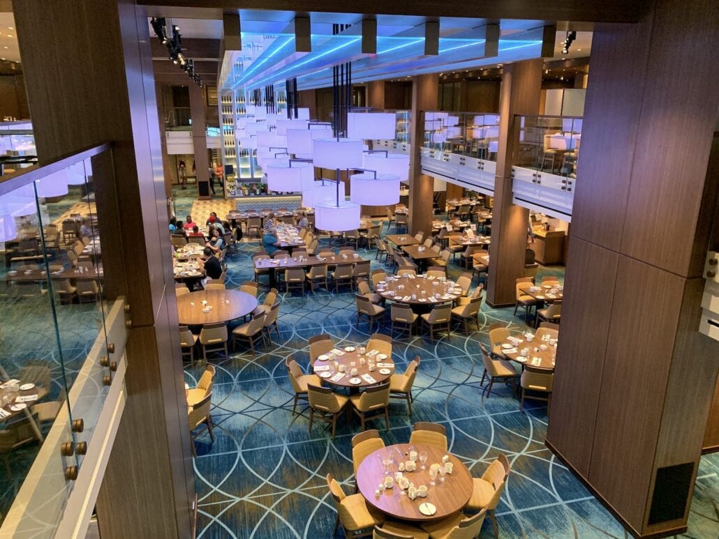 A spacious restaurant dining area aboard the Carnival Vista features multiple round tables set for service. The room, viewed from an elevated position, boasts wooden decor, blue carpeting with a geometric pattern, and modern light fixtures—truly a visual highlight on this cruise experience.