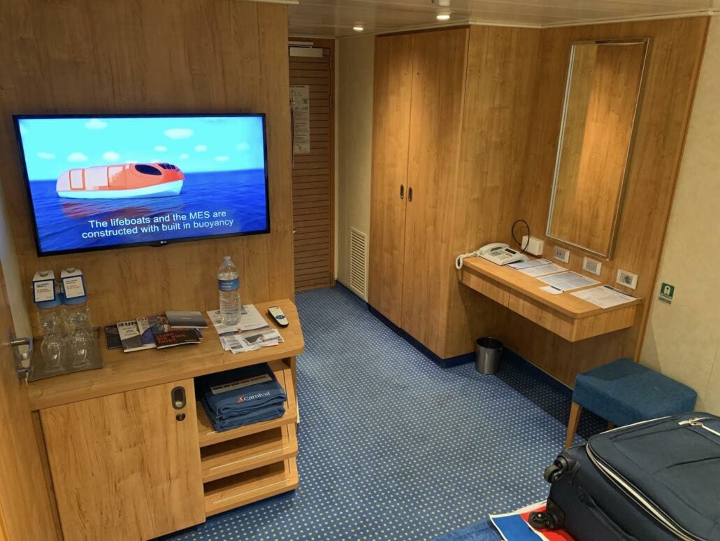 A cruise cabin aboard the Carnival Vista boasts wood-paneled walls and a blue carpet, with a wall-mounted TV playing an animation. It includes a desk with a mirror, closet, small blue ottoman, suitcase, and informational brochures on the bed—perfect for your next cruise review.