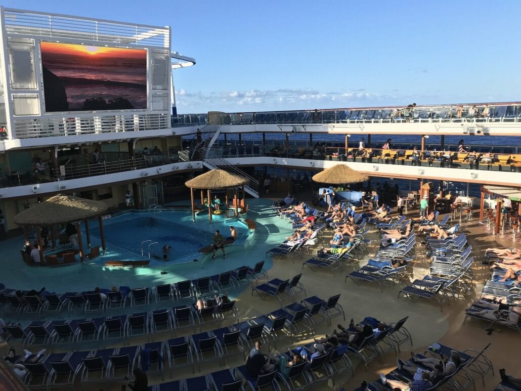 A cruise ship deck on the Carnival Vista with a large outdoor pool surrounded by lounge chairs exudes relaxation. Guests are sunbathing, unwinding beneath a sky with a few clouds, while a giant screen displays a stunning sunset over the ocean, crafting the perfect cruise review moment.