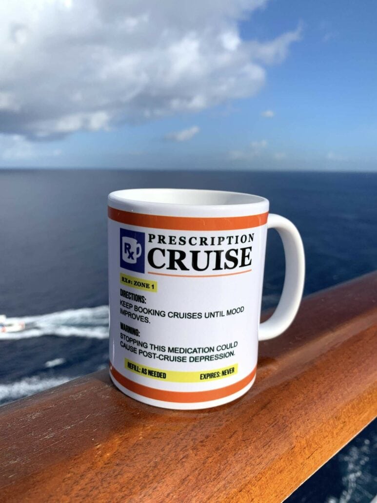 A mug designed like a prescription bottle rests on a wooden railing against a backdrop of ocean and sky. The mug humorously reads "Prescription Cruise," echoing the joy of reading a Carnival Vista cruise review to boost one's mood.