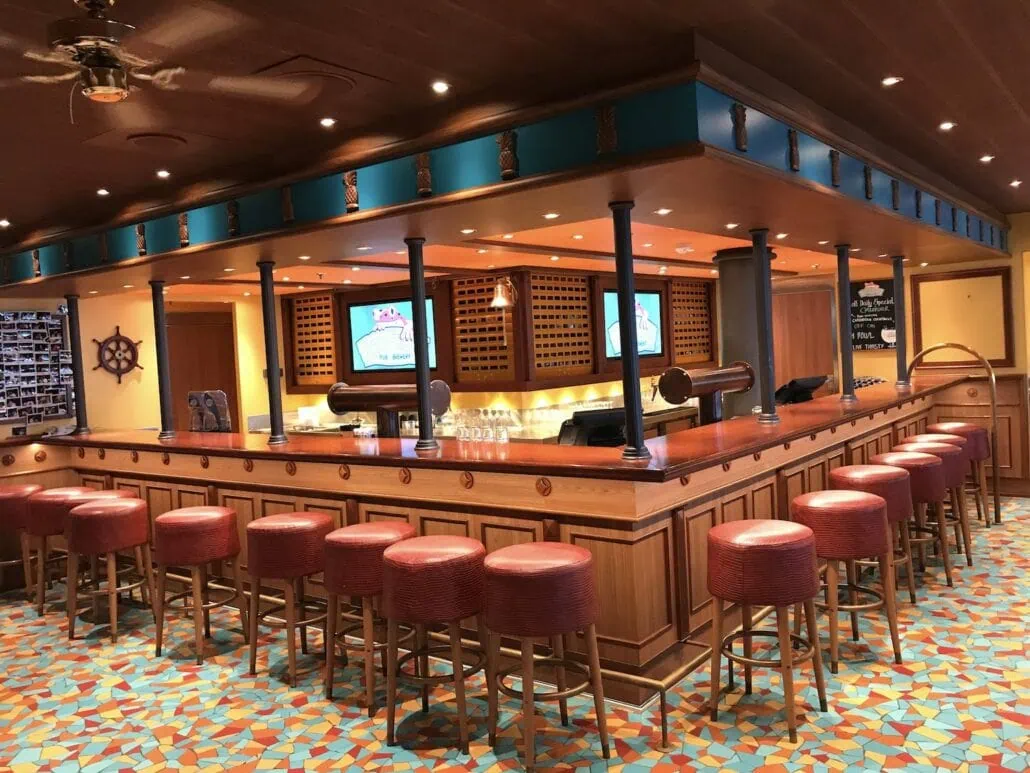 A spacious bar with wooden accents on the Carnival Vista cruise features a long counter surrounded by red cushioned stools. Overhead lights, beverage taps, and wall-mounted screens display drink options. The floor is adorned with a vibrant mosaic pattern, adding to the lively atmosphere.