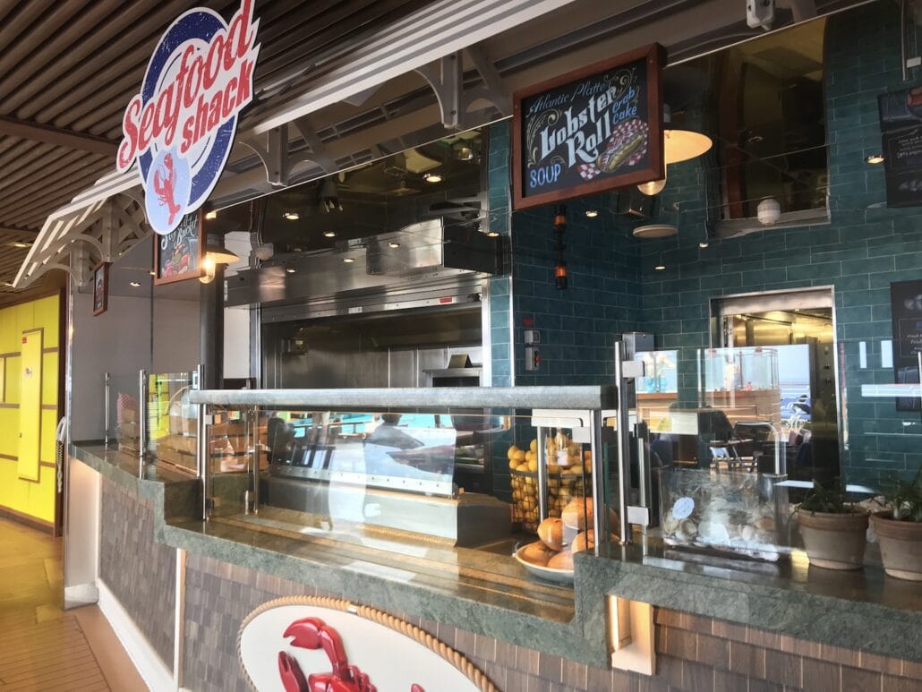 A seafood snack bar aboard the Carnival Vista features a menu sign boasting a lobster roll and soup. Glass displays reveal tempting bites, with kitchen equipment glimpsed in the background. Above, a seafood shack sign nods to oceanic delights enjoyed at sea.