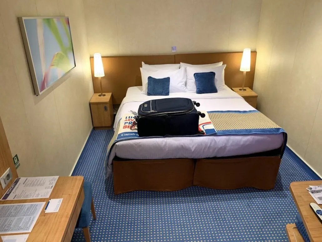 The hotel room, reminiscent of a Carnival Vista cruise review, features a neatly made double bed with two bedside lamps casting a warm glow over the wall painting. Blue carpeting adds charm, while a suitcase rests on the bed. A small desk nearby holds an array of papers and a telephone.
