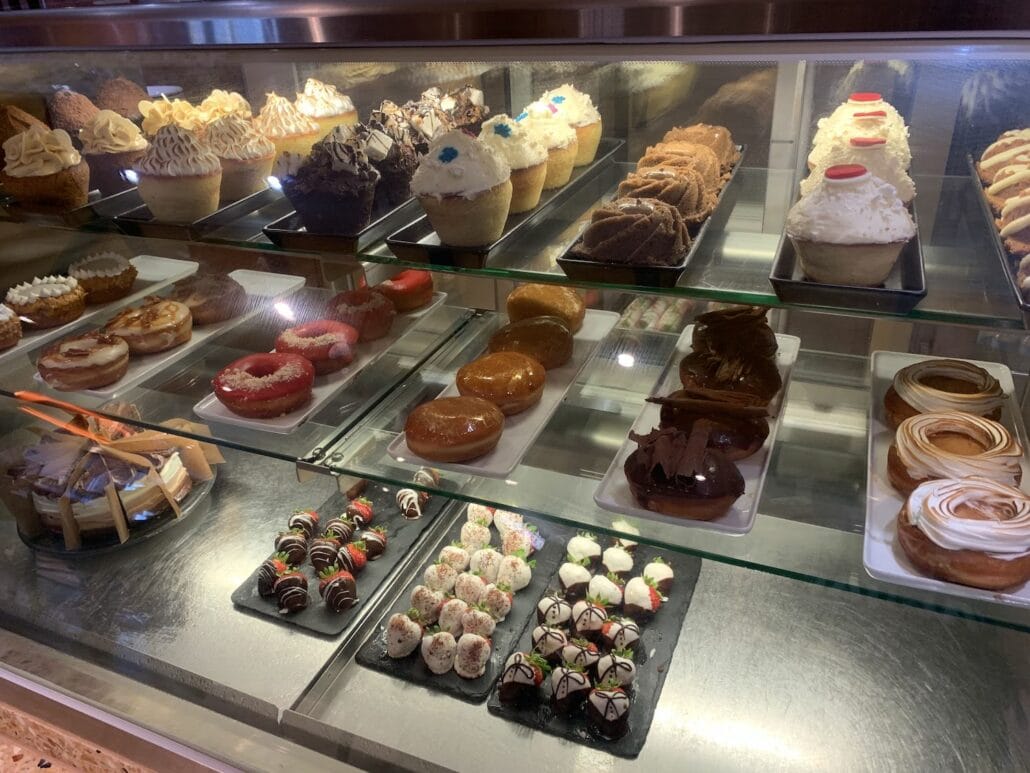A display case filled with various desserts, including cupcakes, pastries, donuts, and chocolates, reminiscent of the delightful offerings found on a Carnival Vista cruise. Each shelf is neatly arranged with different types of sweet treats on plates and trays.