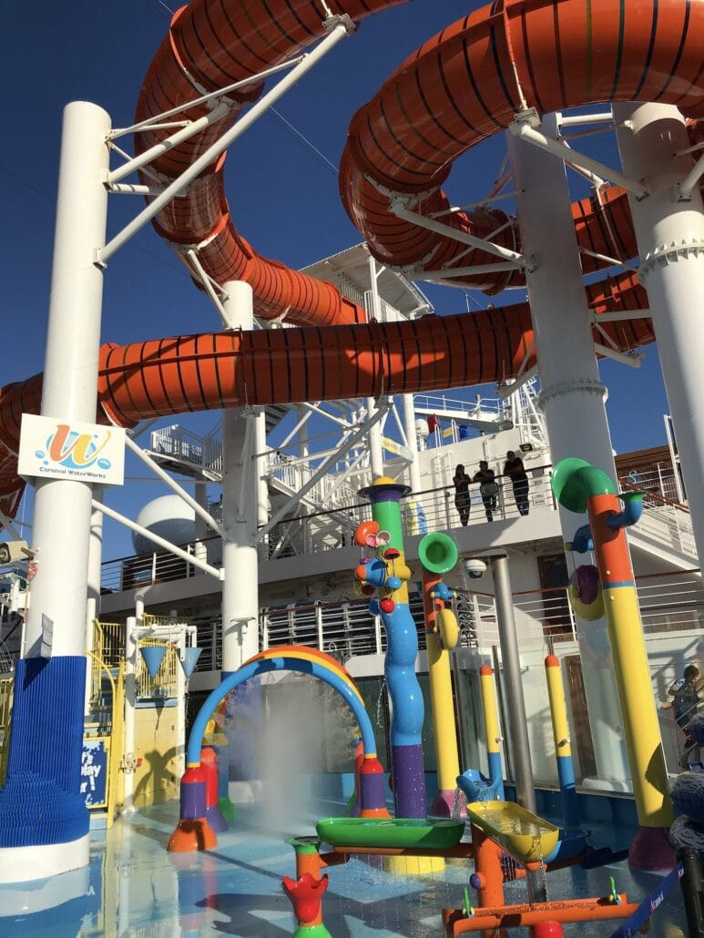 A colorful water park on the Carnival Vista features large, twisting orange slides supported by white columns. Below, playful water structures entertain children with spray jets and splash zones. The clear, blue sky enhances the vibrant atmosphere onboard this cruise experience.