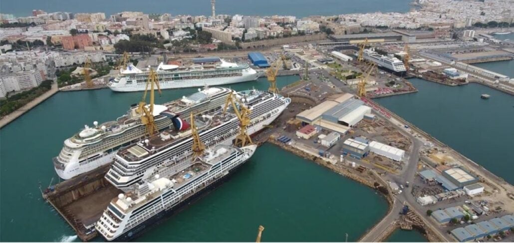 Aerial view of a shipyard with multiple cruise ships, including Carnival Destiny, docked for maintenance, surrounded by cranes and shipyard buildings, near a coastal city.