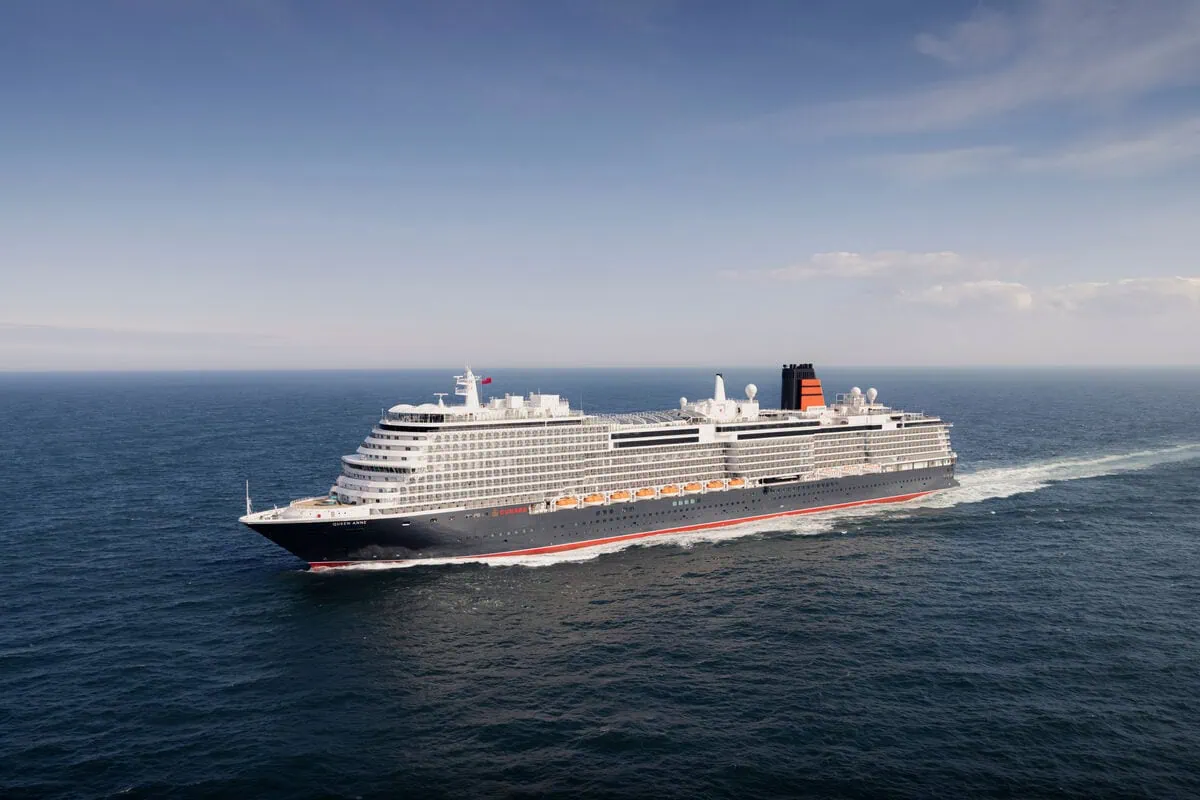 A large Cunard ship sails through a calm ocean under a clear sky. The vessel, painted in dark and light colors, boasts multiple decks and showcases the grandeur that ranges from the smallest to largest in their fleet.