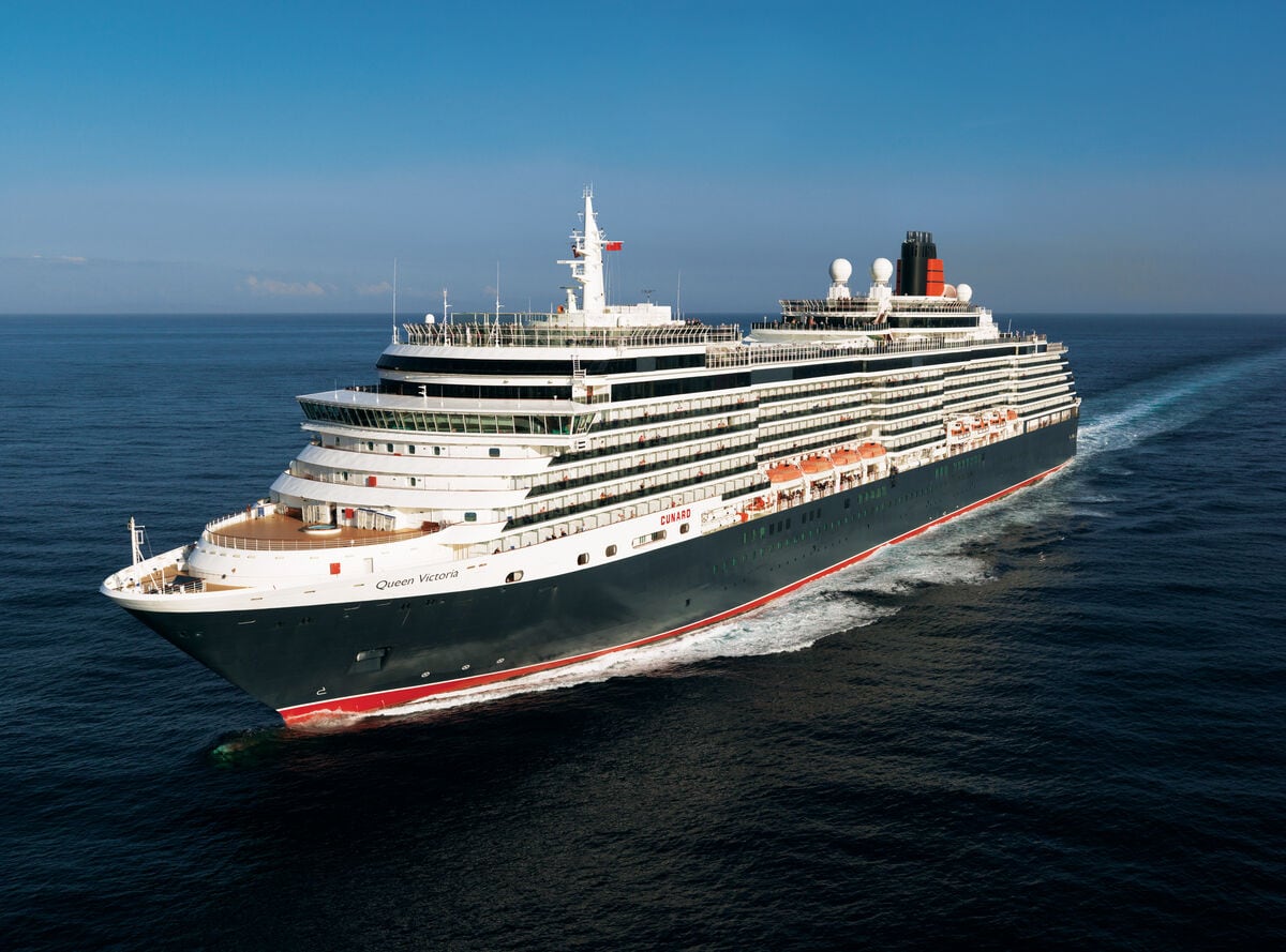 A large Cunard ship, the "Queen Victoria," sails on calm, blue ocean waters under a clear sky.